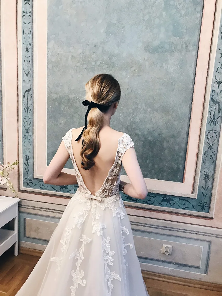 Bride in a lace open-back gown facing an ornate mirror, hair in a curled low ponytail tied with black velvet ribbon