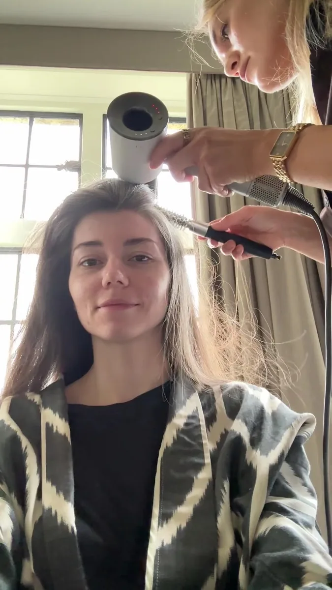 Stylist blow-drying and combing a seated client's long dark hair beside a window