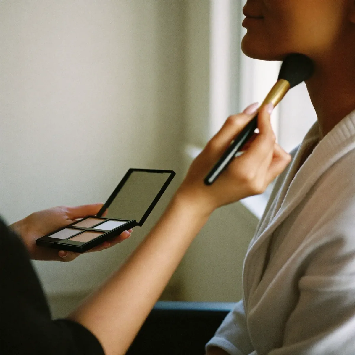 Makeup being applied with a soft brush in a bright getting-ready room